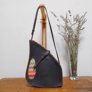 Può includere: Borsa a tracolla in pelle marrone scuro dalla forma a goccia unica. La borsa presenta un inserto in tessuto colorato fantasia e un cinturino in pelle marrone con fibbia. La borsa è appesa a un supporto in legno.