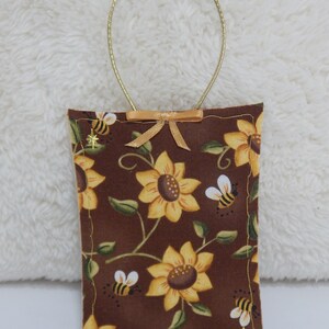 May include: A brown fabric sachet with a gold cord hanger. The sachet is decorated with a sunflower and bee pattern.