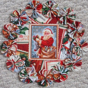 May include: A circular Christmas-themed fabric decoration. The center features a Santa Claus postage stamp design. The border is adorned with fabric rosettes in red, white, and green patterns, creating a festive holiday accent.