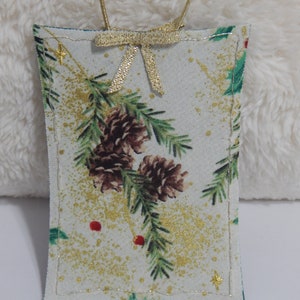 May include: A rectangular Christmas ornament with a festive design. The ornament features a cream-colored background with pine cones, green pine needles, and red berries. Gold glitter accents and a gold bow and string complete the holiday look.