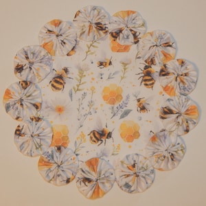 May include: A circular, floral-themed design featuring a central pattern of bees, honeycomb, and flowers. The outer edge is adorned with fabric circles in white, orange, and yellow, with bee and floral patterns. The overall design has a watercolor aesthetic.