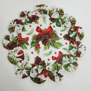 May include: A round, white Christmas placemat with a scalloped edge. The center features pine cones, holly, and red bows. The edge is decorated with fabric circles in a matching pattern. Festive holiday decor.