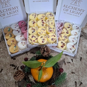 May include: Three boxes of tea light candles with the text "ALCHEMY OF LIGHT MY HOME SCENTS". The candles are yellow, pink, and white, some with dried flower petals. A wreath with a tangerine and pinecones is in the foreground.