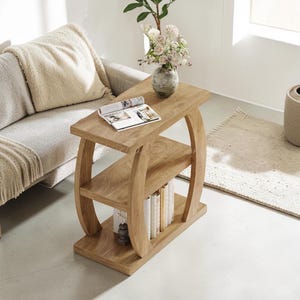 May include: A light brown wooden side table with a rectangular top and a middle shelf, supported by two curved side panels. The table features a wood grain finish and holds an open book on the top surface, with several books stored on the bottom shelf.