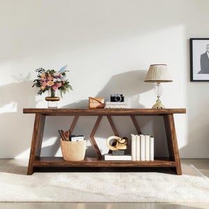 May include: A dark wood console table with a geometric base and a lower shelf. The top holds a glass vase with a floral arrangement, a small brown bag, a camera, and a table lamp. The lower shelf contains a woven basket, a gold sculpture, and books.