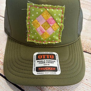 May include: Olive green trucker hat with a patchwork design. The patch features a green polka dot background with pink and yellow squares. The hat has a mesh back and the text "OTTO TRUCKER" on the front.