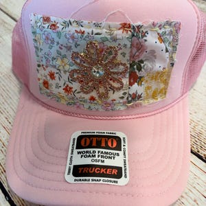 May include: Pink trucker hat with a floral patch and a decorative flower. The hat features a mesh back and a curved brim. The patch has a variety of floral patterns. The hat has the text "OTTO WORLD FAMOUS FOAM FRONT OSFM TRUCKER".