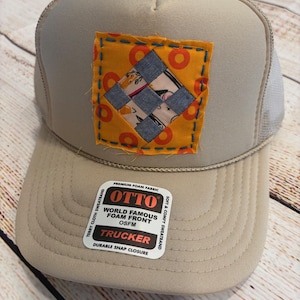 May include: Tan trucker hat with a square patch featuring a patchwork design in orange, blue, and patterned fabrics. The patch is stitched onto the hat with blue thread. The hat has a white mesh back and a snap closure. The hat has a tag that says "OTTO TRUCKER".