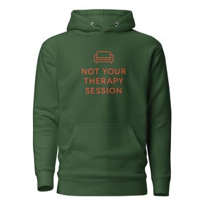Puede incluir: Una sudadera con capucha verde oscuro con bolsillo delantero y cordón. En la parte delantera, el texto naranja "NOT YOUR THERAPY SESSION" con un pequeño gráfico de sofá naranja.