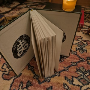 May include: An open book with a dark green cover and a black and white illustration on the inside cover. The pages are white and the book is resting on a patterned rug. A lit candle in a glass is in the background.