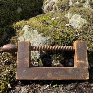 Könnte beinhalten: Eine alte Holzzwinge mit dunkelbrauner Oberfläche, einem rechteckigen Rahmen und einem Gewindeschraubmechanismus mit abgerundetem Griff. Das Werkzeug weist Gebrauchsspuren auf der Holzoberfläche auf und ist horizontal ausgerichtet.