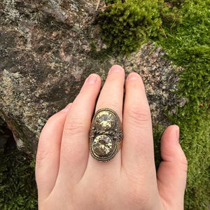 Può includere: Un anello dorato antico con due grandi gemme ovali giallo chiaro. L'anello presenta un bordo dettagliato a forma di corda e un'estetica vintage. L'anello è indossato su una mano, mostrando le sue dimensioni e il suo design.