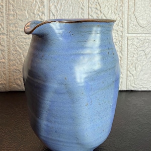 Può includere: Una brocca in ceramica blu fatta a mano con un beccuccio. La brocca ha un corpo arrotondato e un bordo leggermente svasato con un bordo marrone. La glassa blu presenta sottili variazioni di colore, conferendole un aspetto rustico. Questa brocca è adatta per servire bevande.