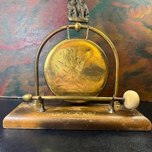 Könnte beinhalten: Ein Vintage-Gong aus Messing mit Holzsockel. Der Gong ist in einem Messingrahmen aufgehängt und hat eine kleine Skulptur von drei Figuren oben. Ein Schlägel mit hellem Kopf ruht auf dem Holzsockel. Der Gong hat eine warme Goldfarbe.