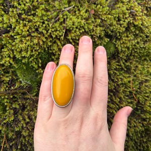 May include: A large, oval ring featuring a bright yellow stone set in a silver-coloured metal band. The ring is shown on a hand against a backdrop of green moss.