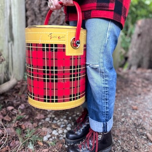 May include: A vintage "Scotch Kooler" cooler with a red and black plaid pattern. The cooler has a yellow top and bottom with a red handle. The text "Scotch Kooler" is visible on the top. The cooler is cylindrical in shape.