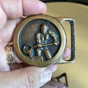 May include: Vintage brass belt buckle featuring a relief of a blacksmith figure. The circular design depicts a man working with a tool, set against a dark background. The buckle has a rectangular frame and a tarnished, aged appearance.