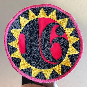 May include: Embroidered patch with a circular design. The patch features a red center with the number "16" in black. A ring of yellow triangles surrounds the number, set against a black background. The outer edge is pink.