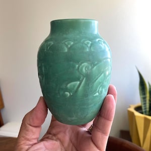 May include: A small, green vase with a matte finish. The vase features a detailed, raised floral pattern around its body. The vase is held in a hand, showcasing its size and shape. The vase is a decorative item.