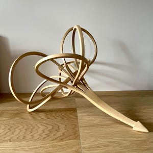 May include: A light-colored wooden sculpture with a complex, intertwined design. The sculpture features multiple circular and curved elements, with a long, pointed base. The piece is displayed on a wooden surface.