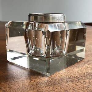 Könnte beinhalten: Ein klarer, facettierter Glas-Tintenbehälter mit silberfarbenem Deckel. Der Tintenbehälter hat eine quadratische Basis und ein zylindrisches Tintenreservoir. Der Deckel ist mit "STERLING" gekennzeichnet. Ein klassisches Schreibtischaccessoire.