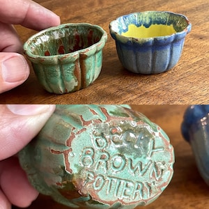 May include: Two small, fluted ceramic bowls with scalloped edges. One bowl features a green and brown glaze, while the other has a blue and yellow glaze. The underside of the green bowl is embossed with the text "BECKY BROWN POTTERY" in raised letters.