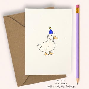 Puede incluir: Una tarjeta de felicitación blanca con una ilustración dibujada a mano de un pato que lleva un gorro de fiesta azul con un pompón amarillo. La tarjeta es de tamaño A7, mide 74 x 105 mm, y viene con un sobre marrón y un lápiz morado.