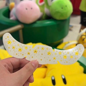 以下が含まれることがあります： 目の下に貼るための湾曲した白いアイパッチ。表面には黒い縁取りの小さな黄色い星と、散りばめられた小さな金色のグリッタードットが繰り返し模様としてデザインされており、目の下の皮膚に貼付するように作られています。