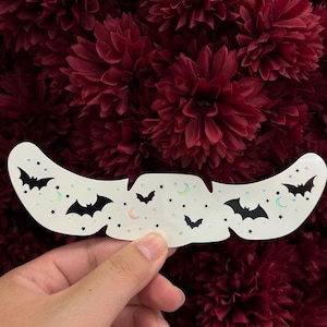 May include: A white, curved nose pore strip featuring a repeating pattern of black silhouettes of flying bats, small iridescent crescent moons, and tiny stars scattered across the surface of the adhesive material.