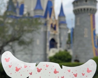 May include: A white, curved adhesive strip featuring a repeating pattern of small, pink, glittery mouse-ear silhouettes and tiny stars. The strip has a scalloped edge design and is held against a blurred background of a large, blue-roofed castle.