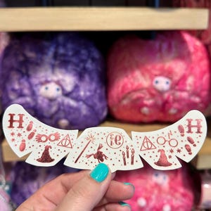 May include: A white, curved adhesive patch featuring a repeating pattern of metallic red glitter icons including the letter H, a wizard hat, round glasses, a wand, a lightning bolt, a deathly hallows symbol, candles, and the number 9 3/4.