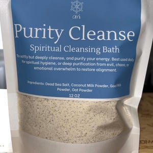 May include: A white pouch containing 12 oz of Purity Cleanse Spiritual Cleansing Bath powder. The blue label features a ship wheel logo, the text House of Omi, and lists ingredients including Dead Sea Salt, Coconut Milk Powder, Goat Milk Powder, and Oat Powder.