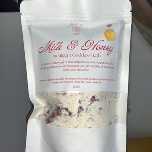 May include: A white bag of Milk & Honey Indulgent Goddess Bath. The bag features text and an illustration of a honey pot with a dripping spoon. The bath product contains goat milk powder, Himalayan pink salt, rosebuds, and rose petals. The bag is labeled 12 OZ.