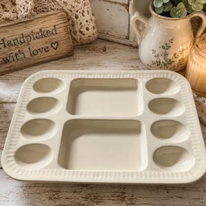 May include: A rectangular cream-colored ceramic serving tray featuring two central square compartments and eight smaller oval-shaped indentations along the sides, finished with a decorative ribbed border around the entire outer edge.