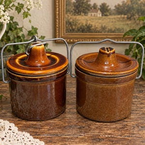May include: Two cylindrical stoneware crocks with glossy brown glaze, each featuring a matching lid with a central knob and a hinged metal wire handle. The crocks have a smooth, uniform finish and are positioned side by side on a wooden surface.