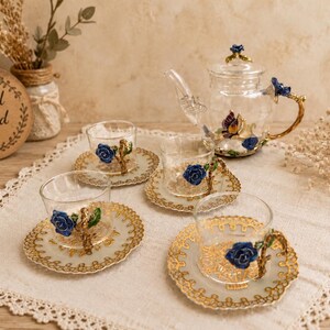 Puede incluir: Un juego de té de cristal transparente que incluye una tetera y cuatro tazas con sus platos a juego. Cada pieza está decorada con detalles en metal dorado, rosas de esmalte azul y motivos de mariposas. La tetera tiene una tapa con una rosa.
