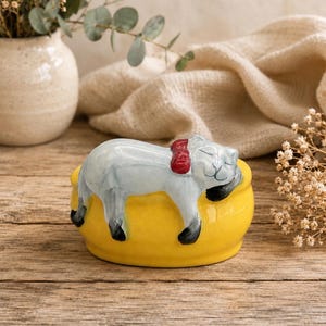 May include: A ceramic cat figurine, painted in shades of gray, white, and black, rests on a bright yellow bowl. The cat wears a red collar. The figurine is set against a rustic wooden surface, with a vase of flowers in the background.