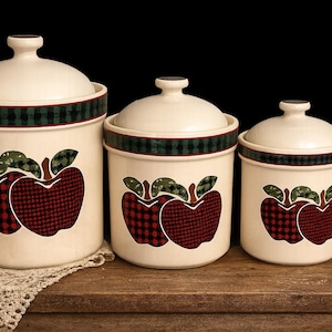 Può includere: Set di tre barattoli in ceramica con coperchi. I barattoli bianco sporco hanno una fascia a quadri verdi e neri e un design a mela a quadri rossi con foglie verdi. Sono esposti su una superficie di legno, offrendo un'estetica rustica.