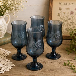 Puede incluir: Cuatro copas de color azul marino con un diseño floral grabado en la superficie. Las copas están dispuestas sobre una mesa de madera, con un jarrón de flores y una impresión enmarcada en el fondo. Las copas miden unos 15 cm de altura.