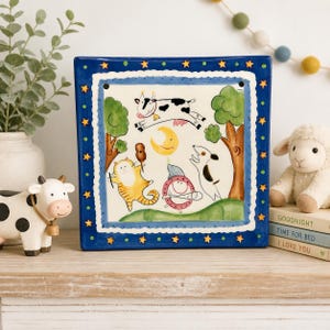 May include: A square, hand-painted ceramic tile featuring a whimsical farm animal scene. The navy blue border has gold stars. The artwork depicts a cow, cat, dog, and moon. The tile is displayed on a wooden surface.
