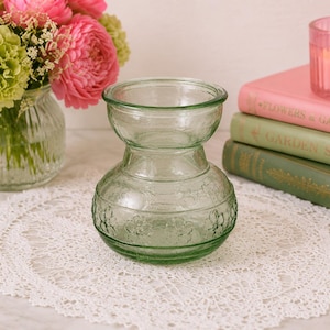 May include: A clear, green glass vase with a bulbous base and a smaller top section. The vase sits on a white lace doily, with a bouquet of pink and green flowers in a similar vase in the background. A stack of books and a lit candle are also visible.
