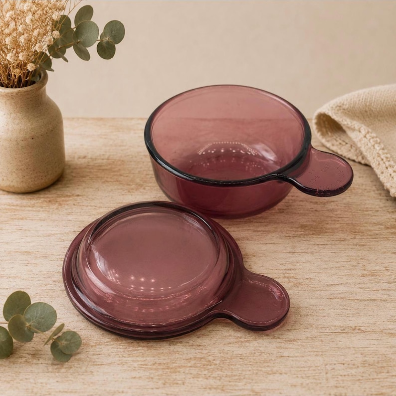 May include: A translucent plum-colored glass bowl with a matching lid, both featuring a small side handle. The bowl is circular with a smooth finish, and the lid is designed to fit securely on top, showcasing a deep purple hue in the glass.