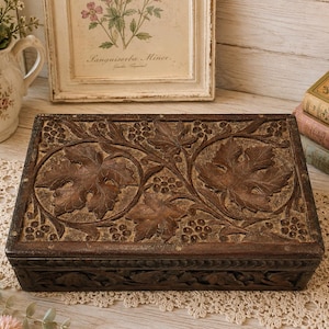 May include: A rectangular dark brown wooden box featuring intricate hand-carved details on the lid and sides. The lid displays a prominent relief pattern of large grape leaves and clusters of grapes, with a matching carved border along the base.