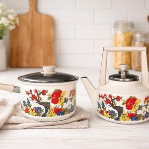 Puede incluir: Juego de cacerola y tetera de esmalte color crema. Ambas piezas presentan un diseño floral con flores rojas, amarillas y azules sobre un fondo blanco. La cacerola tiene una tapa negra y un asa de madera.