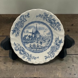 May include: A white ceramic plate with a blue and white windmill and floral design. The plate has a scalloped edge and is supported by a black stand. The plate's centre features a detailed landscape scene.