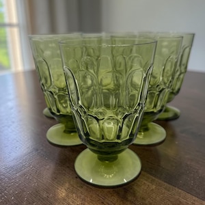 May include: A set of six green-tinted, stemmed drinking glasses. The glasses have a textured, geometric pattern and a rounded base. The glasses are arranged on a dark wooden surface, with natural light in the background.