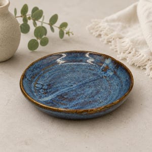 Puede incluir: Un plato redondo de cerámica con un esmalte azul profundo y tonos moteados en marrón y oscuro. La superficie tiene un acabado texturizado e irregular con un relieve visible en el centro y el borde exterior acabado en tono marrón oscuro.