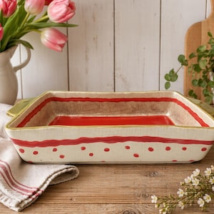 May include: Rectangular ceramic baking dish with a cream base, red accents, and green handles. The dish features a red dotted pattern and a red stripe near the rim. The interior is red. A decorative kitchenware item.