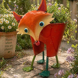 Puede incluir: Una maceta con forma de zorro, pintada en naranja, crema y rojo, con patas y pies verdes. El zorro tiene ojos azules y está lleno de plantas y flores verdes. Un adorno de jardín encantador, perfecto para añadir carácter a los espacios exteriores.