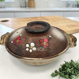 May include: A brown ceramic Japanese donabe pot with a lid. The lid is decorated with red and white floral designs. The pot has a black handle and gold-colored side handles. The pot is sitting on a white surface.